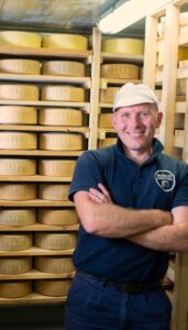 Cheesemaker, Kym Masters, standing in aging room in front of tiers of cheeses.