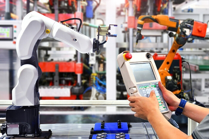 Hands holding a control panel for manufacturing robotics in the background.
